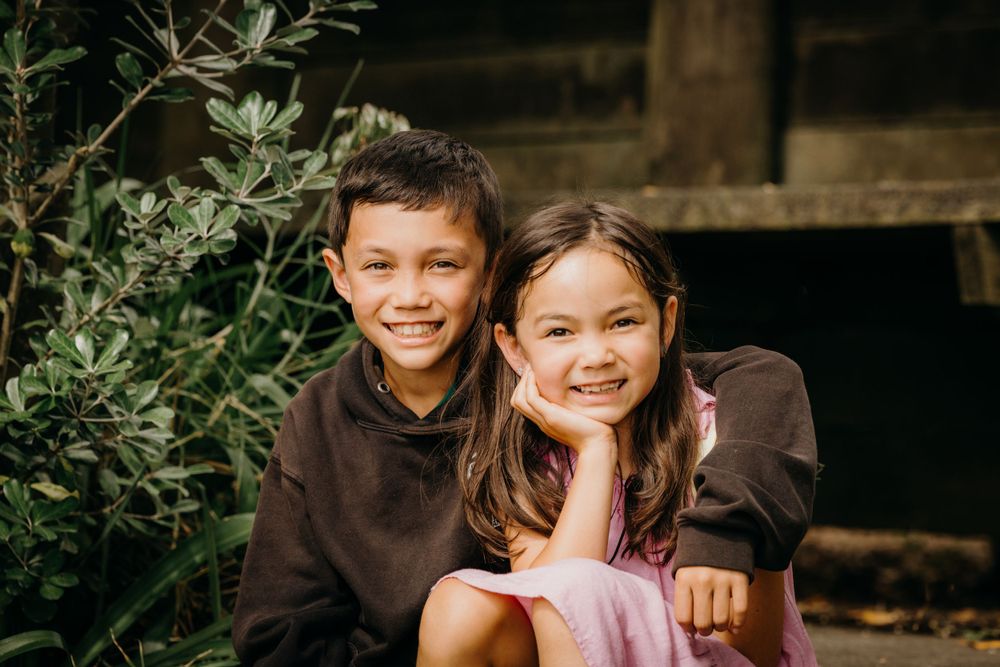 Two kids in brown clothing sit outdoors smiling together beside green foliage.