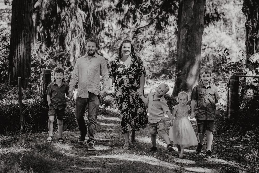 A family walks together on a scenic forest path during a black and white photo session on a sunny day.