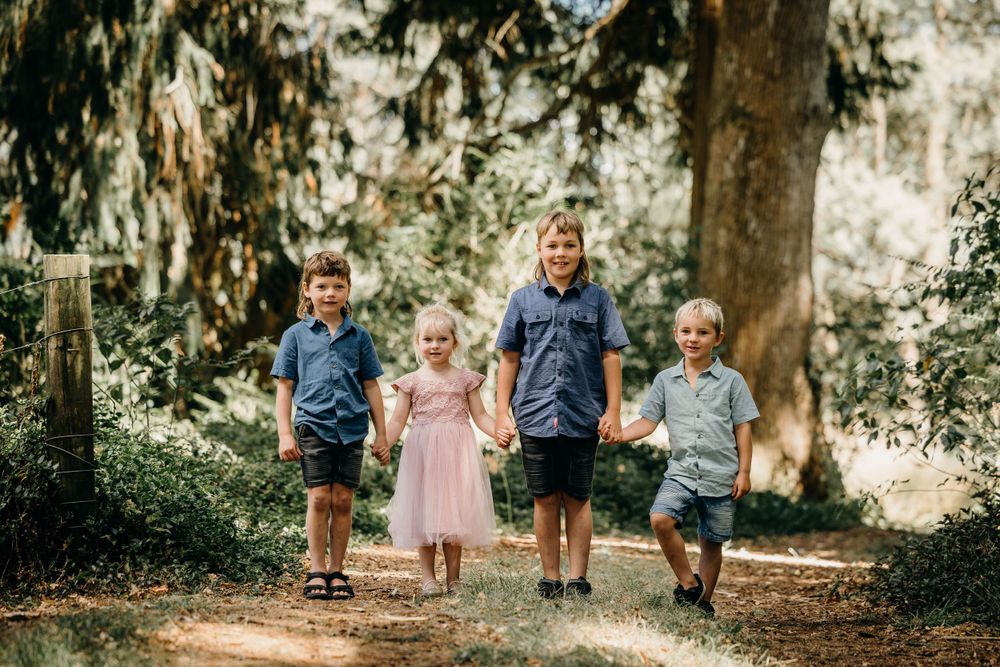 Children in coordinating blue and pink outfits stand together on a nature path surrounded by tall trees in sunlight.