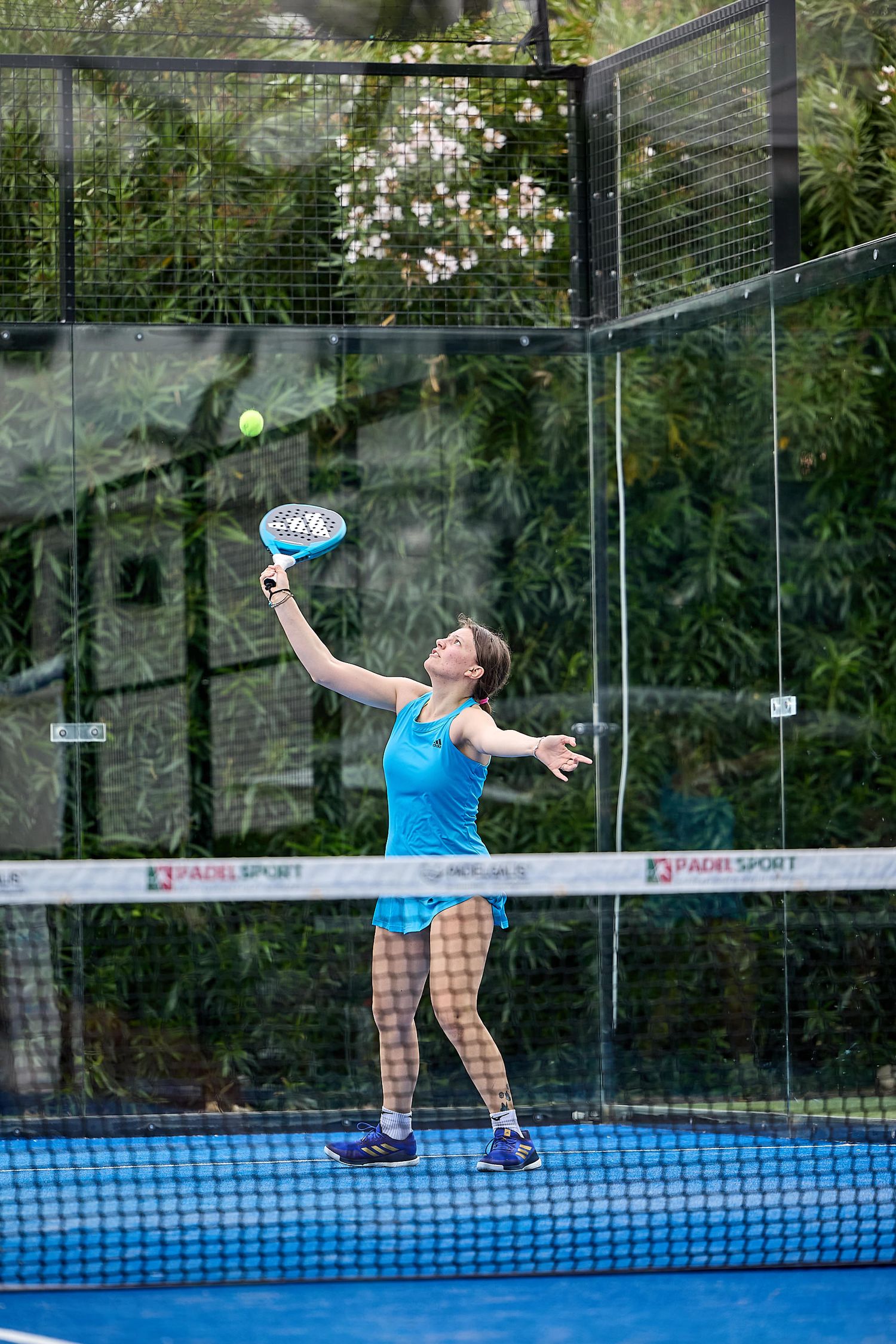 Padel player reaching up to hit overhead shot on blue court surrounded by glass walls.