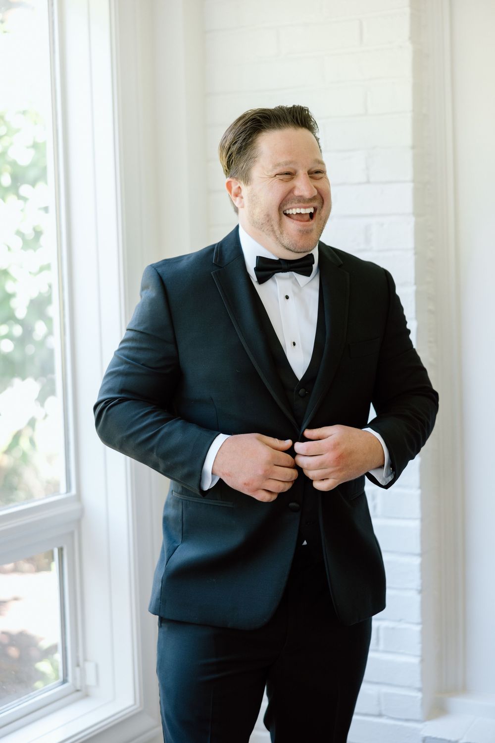 A person wearing a classic black tuxedo laughs joyfully while adjusting their jacket by a bright window.