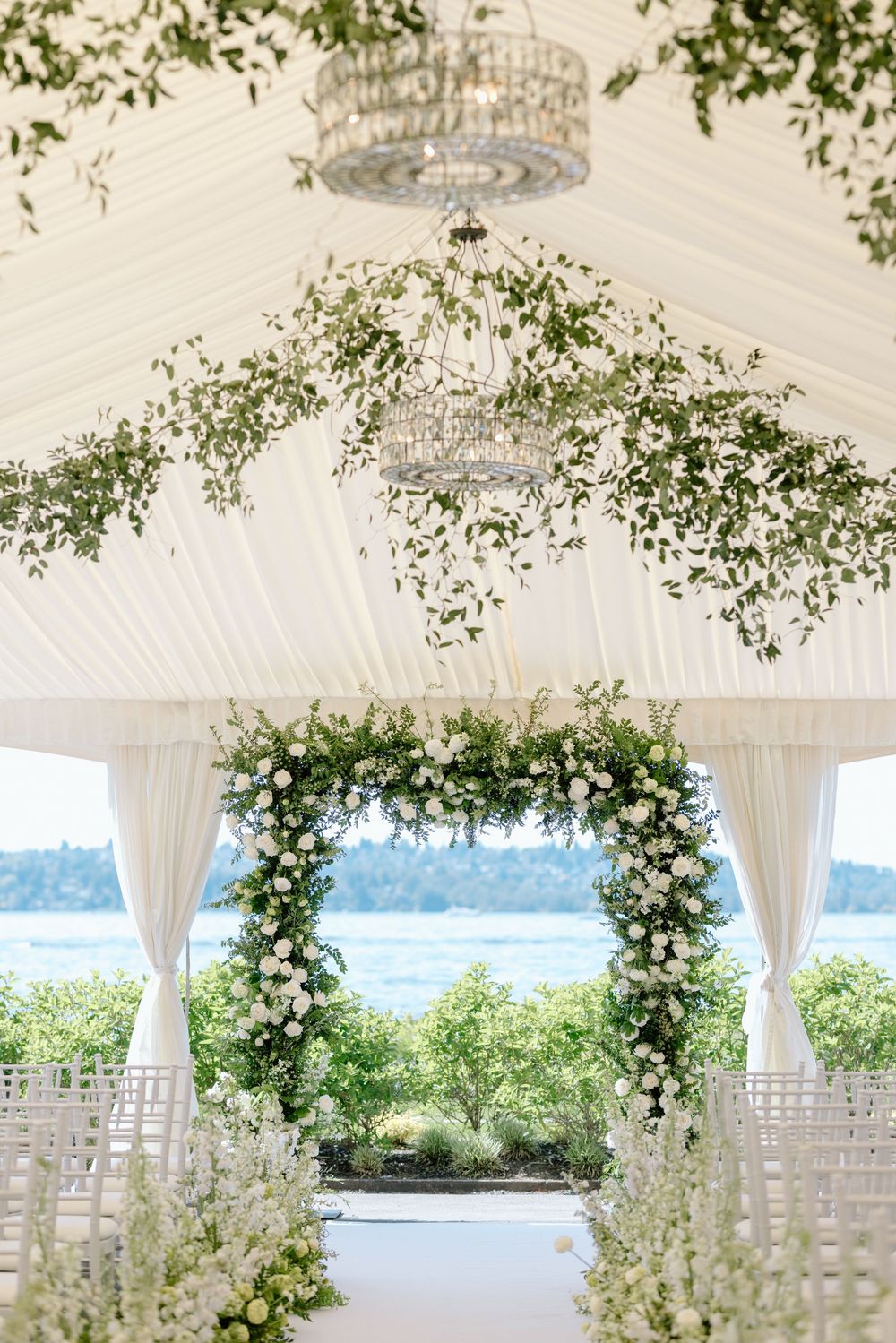 Elegant outdoor wedding ceremony setup with white drapes, chandelier, and floral arch overlooking a waterfront view.