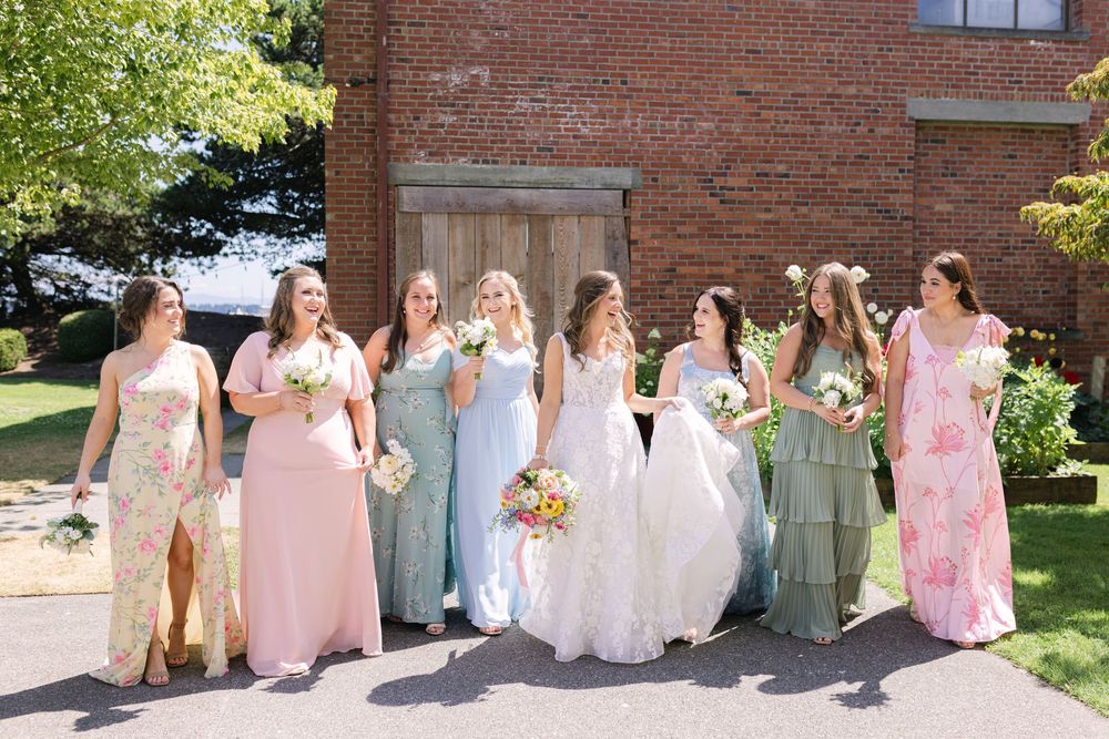 A wedding party in pastel-colored dresses poses outdoors against a brick wall on a sunny spring day.