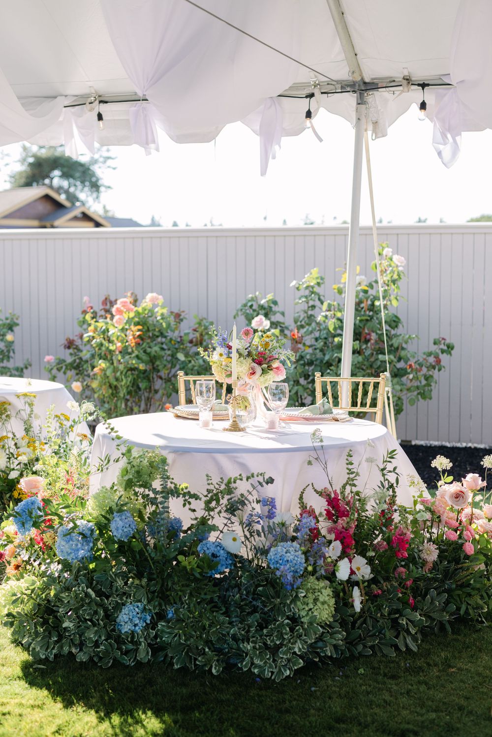 Elegant garden party setting with white table under tent canopy surrounded by blooming hydrangeas and colorful flowers.