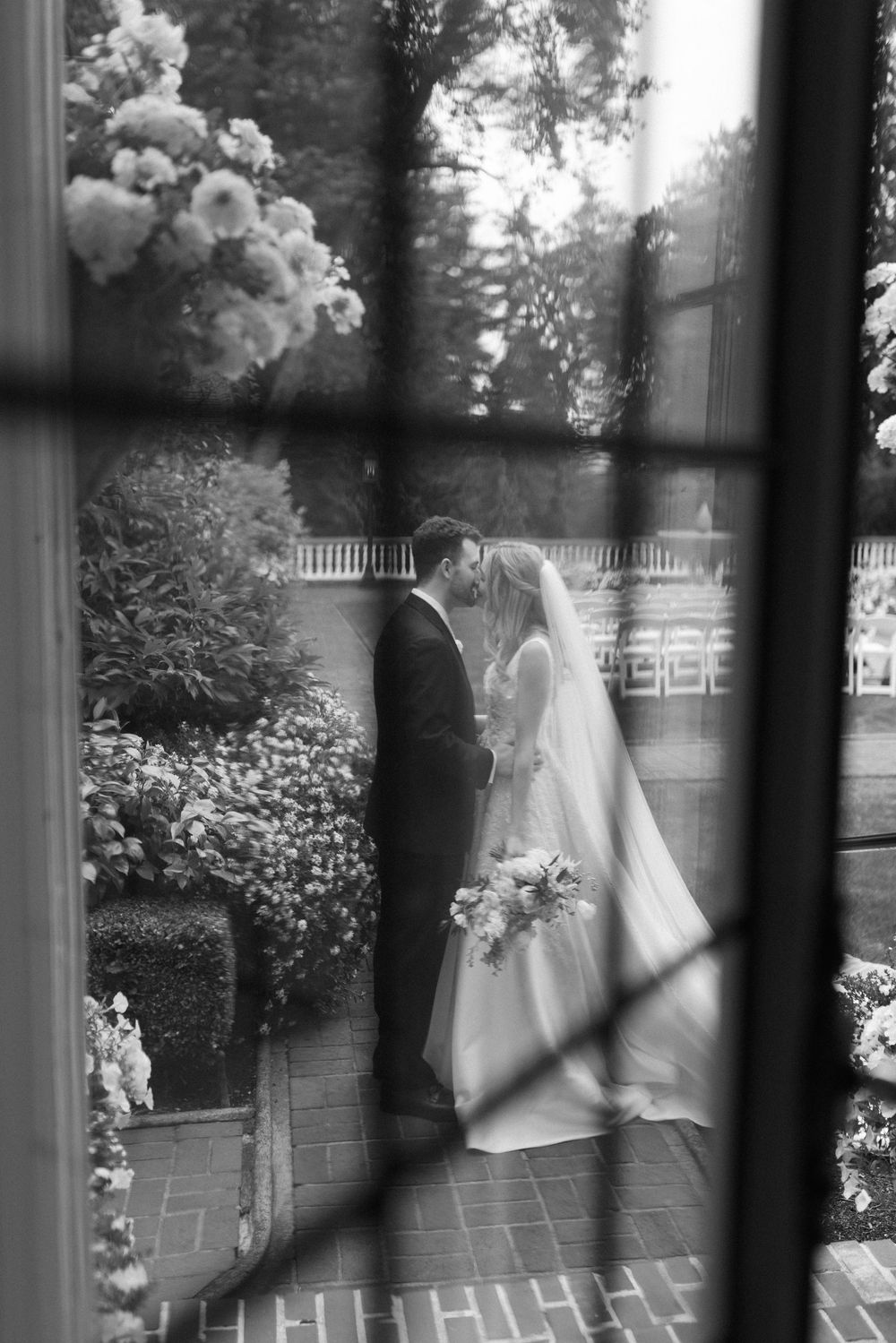 A romantic black and white wedding photo taken through a window showing a couple in a garden courtyard setting.