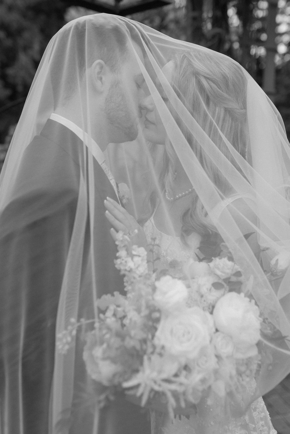A romantic black and white wedding photo showing a couple sharing an intimate moment under a delicate veil with floral bouquet.