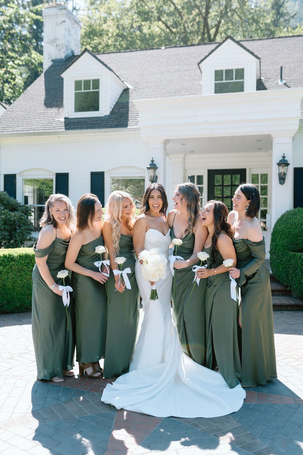 A bridal party poses in sage green bridesmaid dresses and a white wedding gown outside a white colonial-style house.