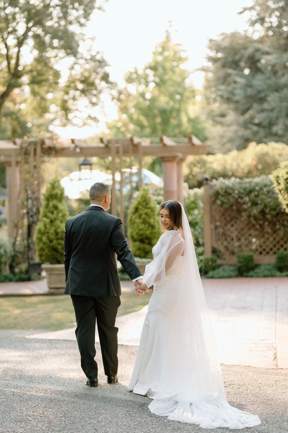 A newlywed couple walks together on a garden path in elegant wedding attire during their outdoor ceremony.