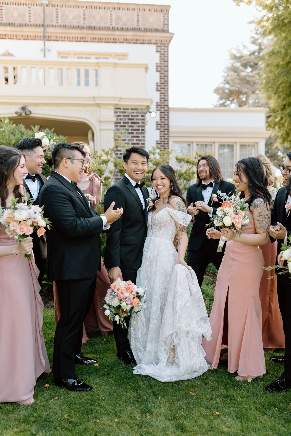 Wedding party in black tuxedos and blush pink bridesmaid dresses laughing together on grass outside elegant venue.