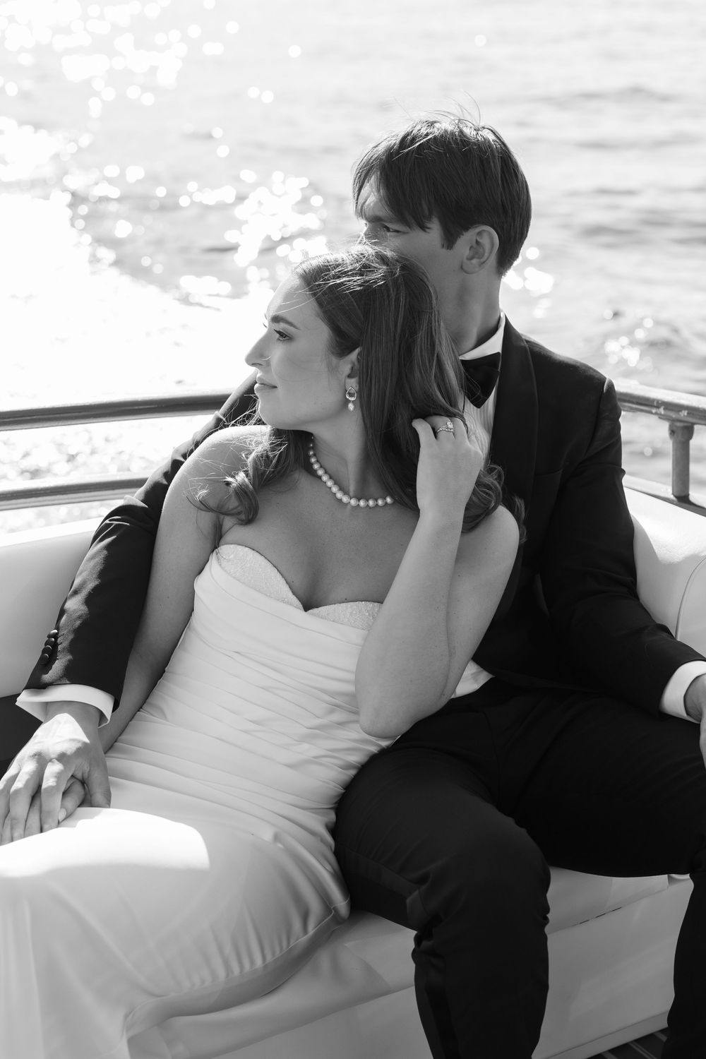 A romantic black and white photograph of a couple in wedding attire sitting together on a boat in sunlit water.