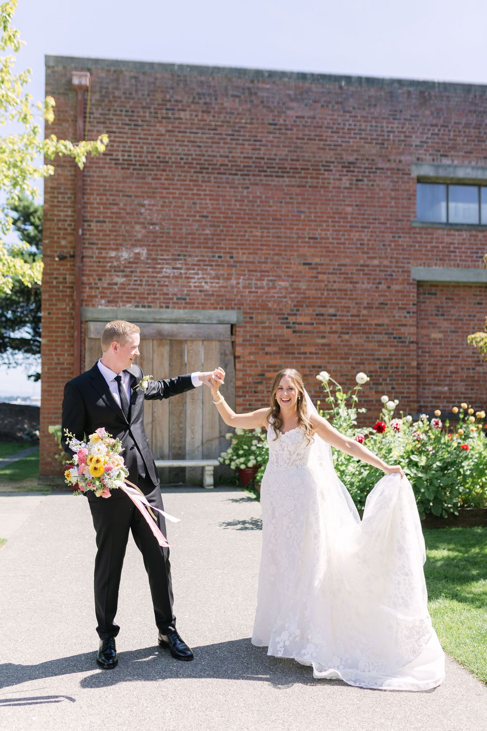 A couple in wedding attire dances together outside a brick building with a flower garden in the background.