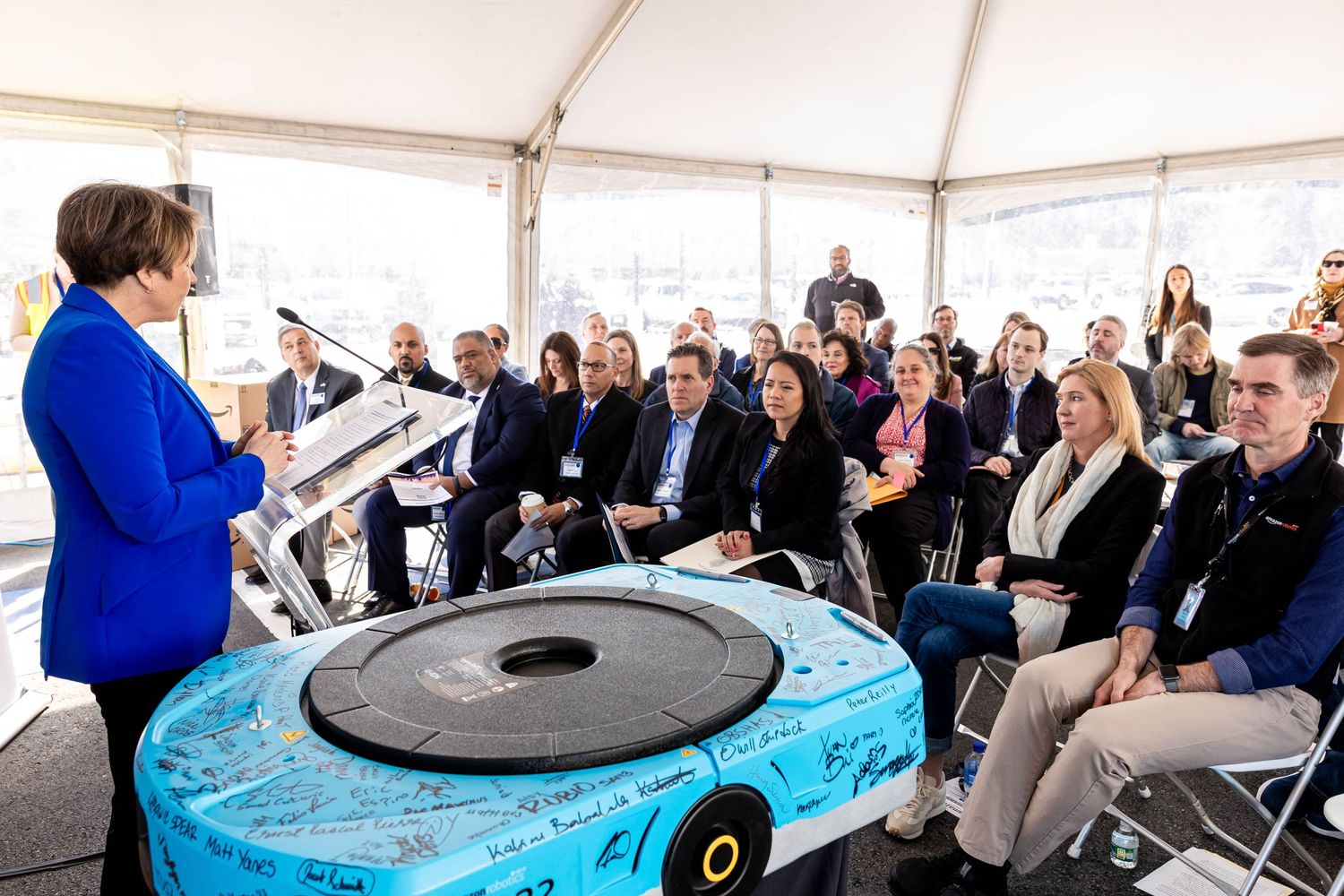 Audience listening to speaker in front of large robotic device in a white tent setting.