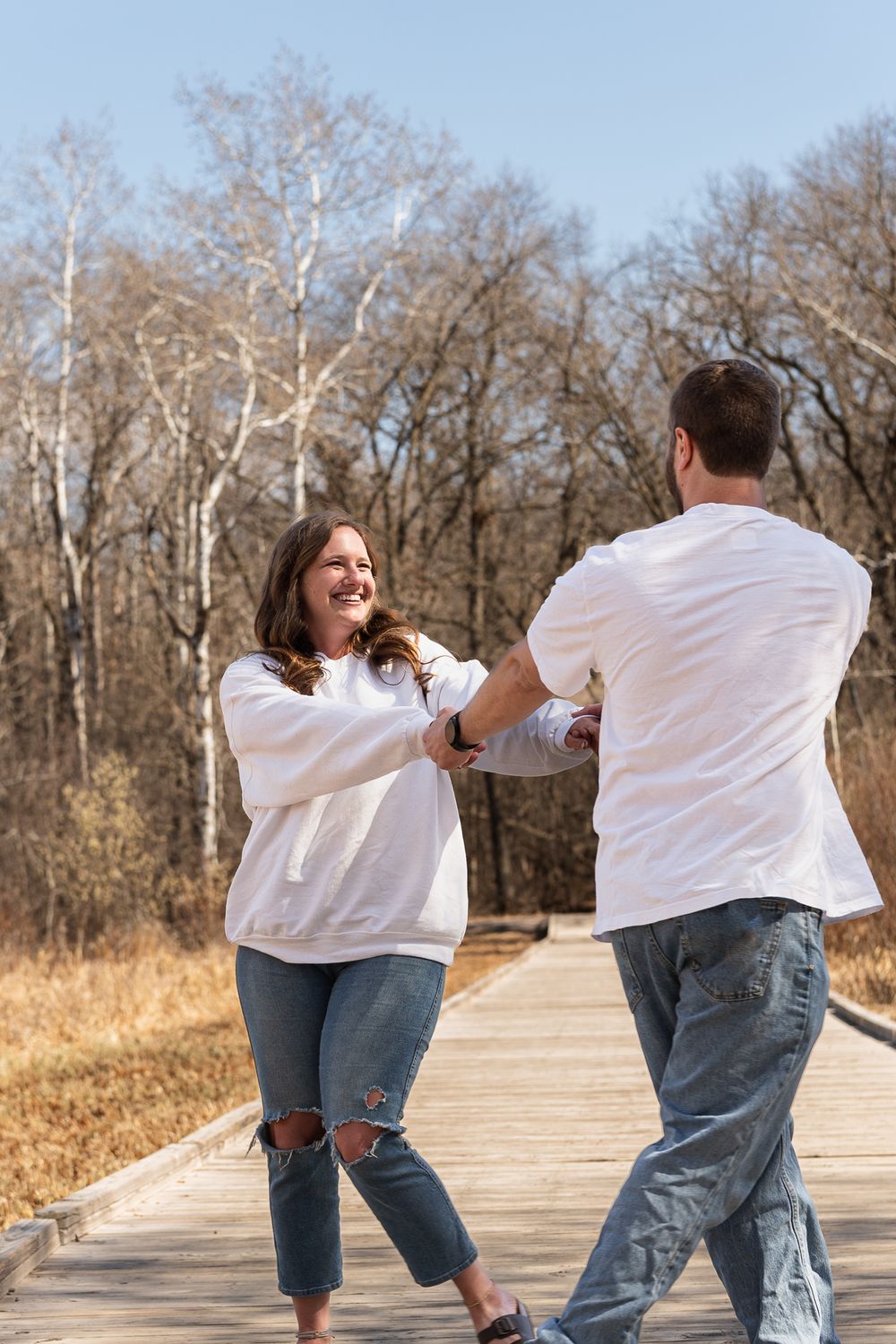 A couple sharing a joyful dance moment on a wooden pathway in casual white outfits.