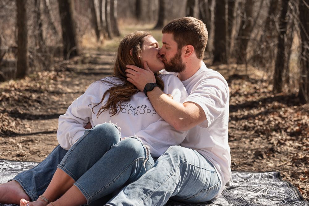 Couple shares intimate moment while sitting on blanket in wooded area.