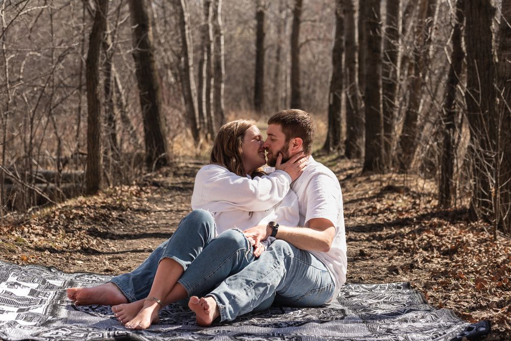 A couple shares an intimate moment while sitting on a blanket in a wooded forest setting.