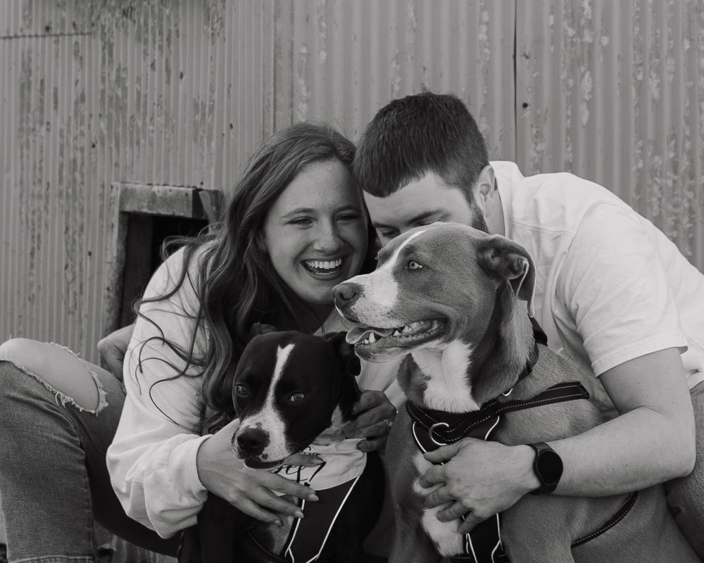 Black and white portrait of couple cuddling with their two dogs outdoors.