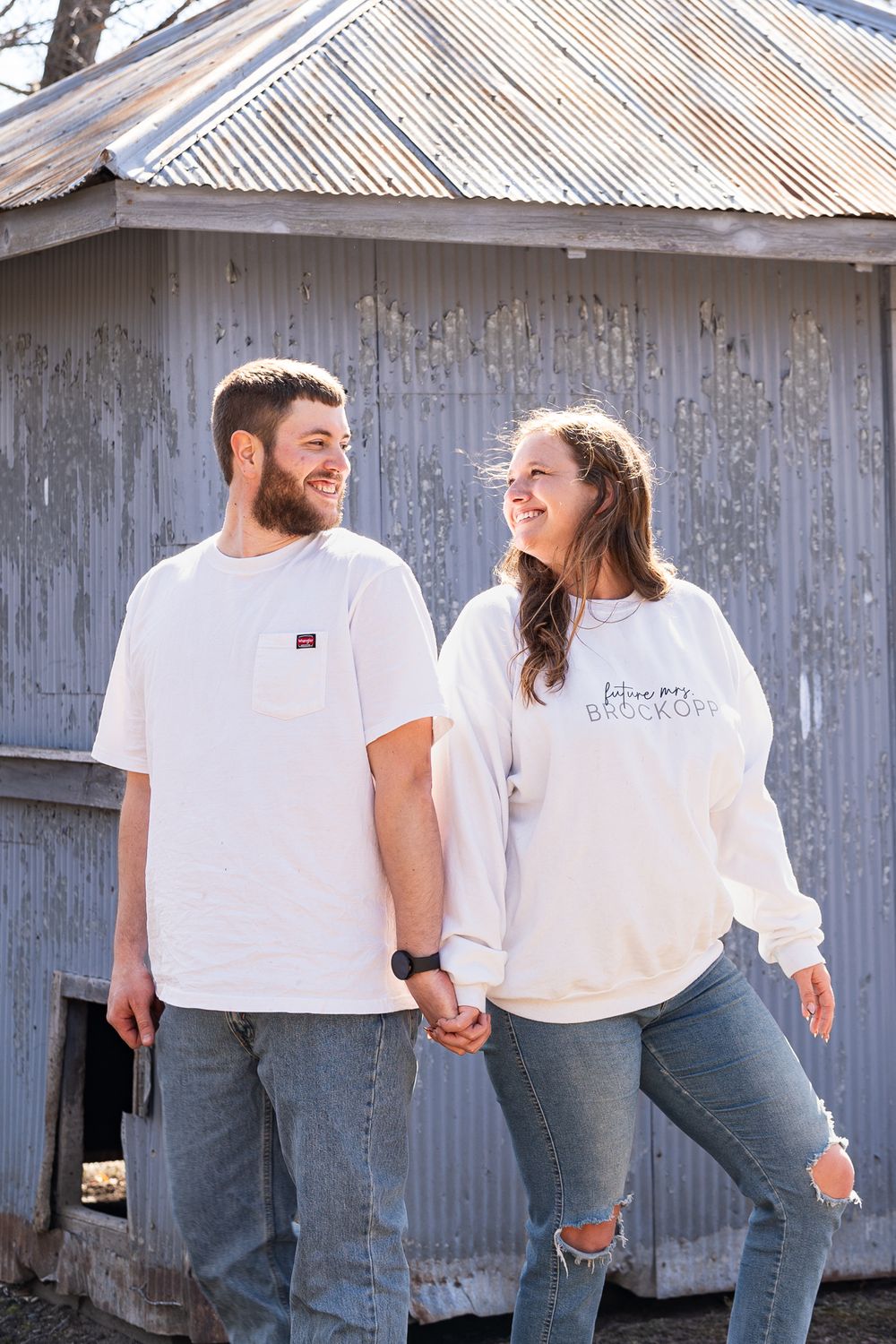 Couple in white outfits holding hands while standing by rustic building.