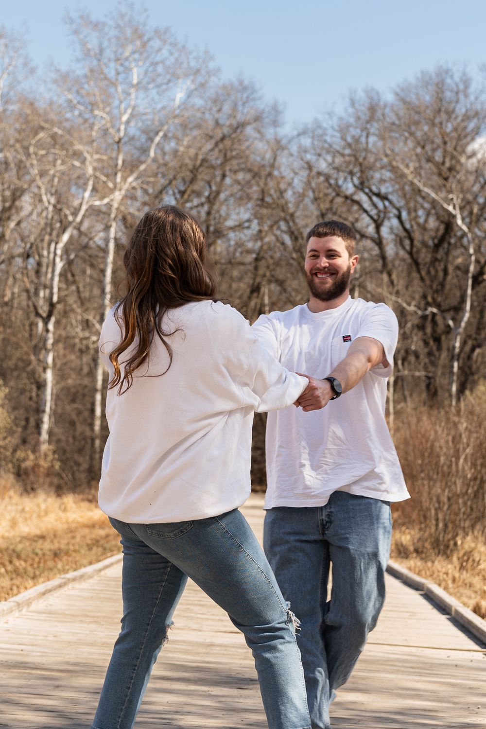 Couple in white shirts dances playfully on a wooden boardwalk surrounded by autumn trees.