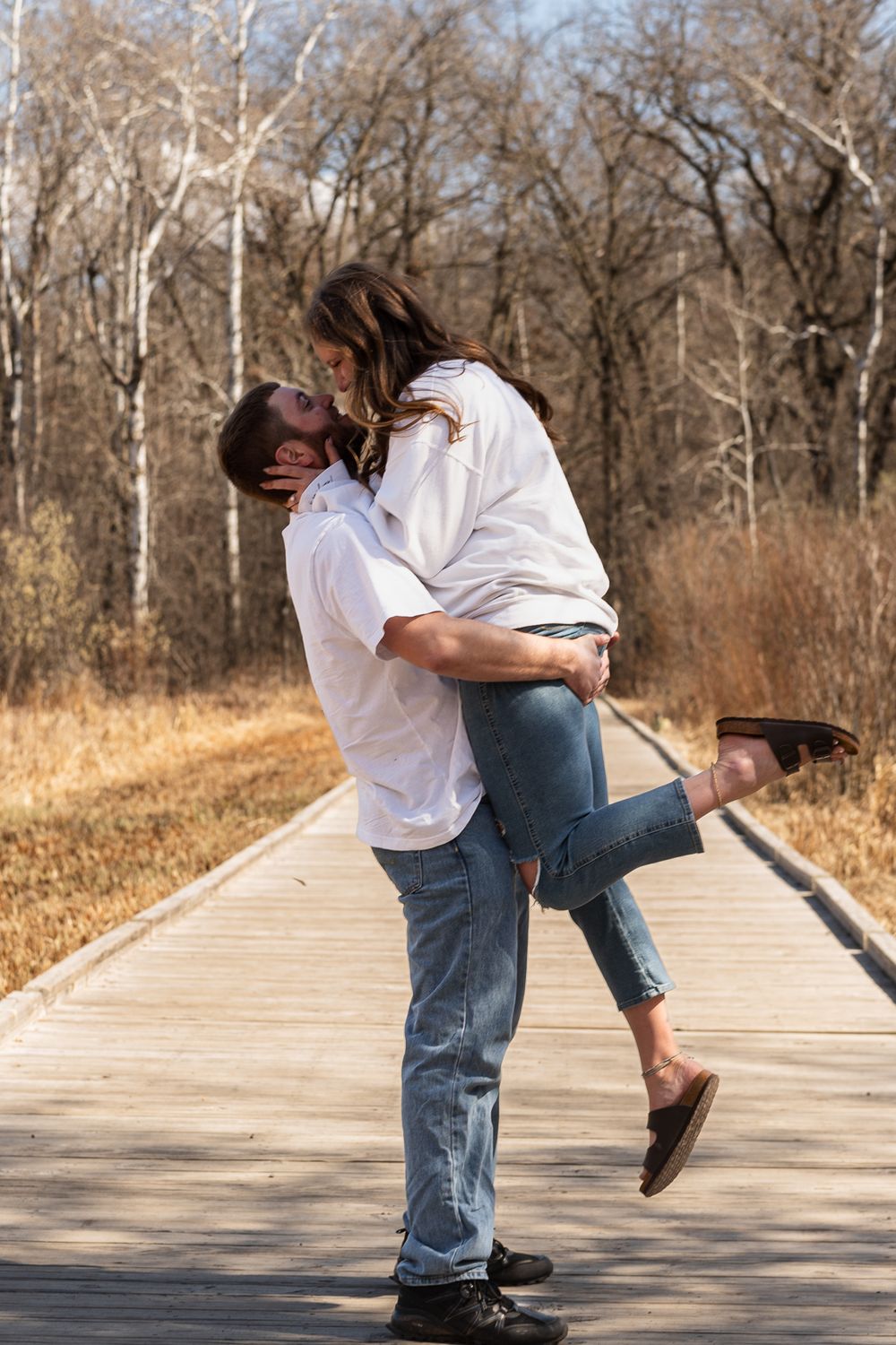 Playful couple in matching outfits embrace on wooden boardwalk during engagement photoshoot.