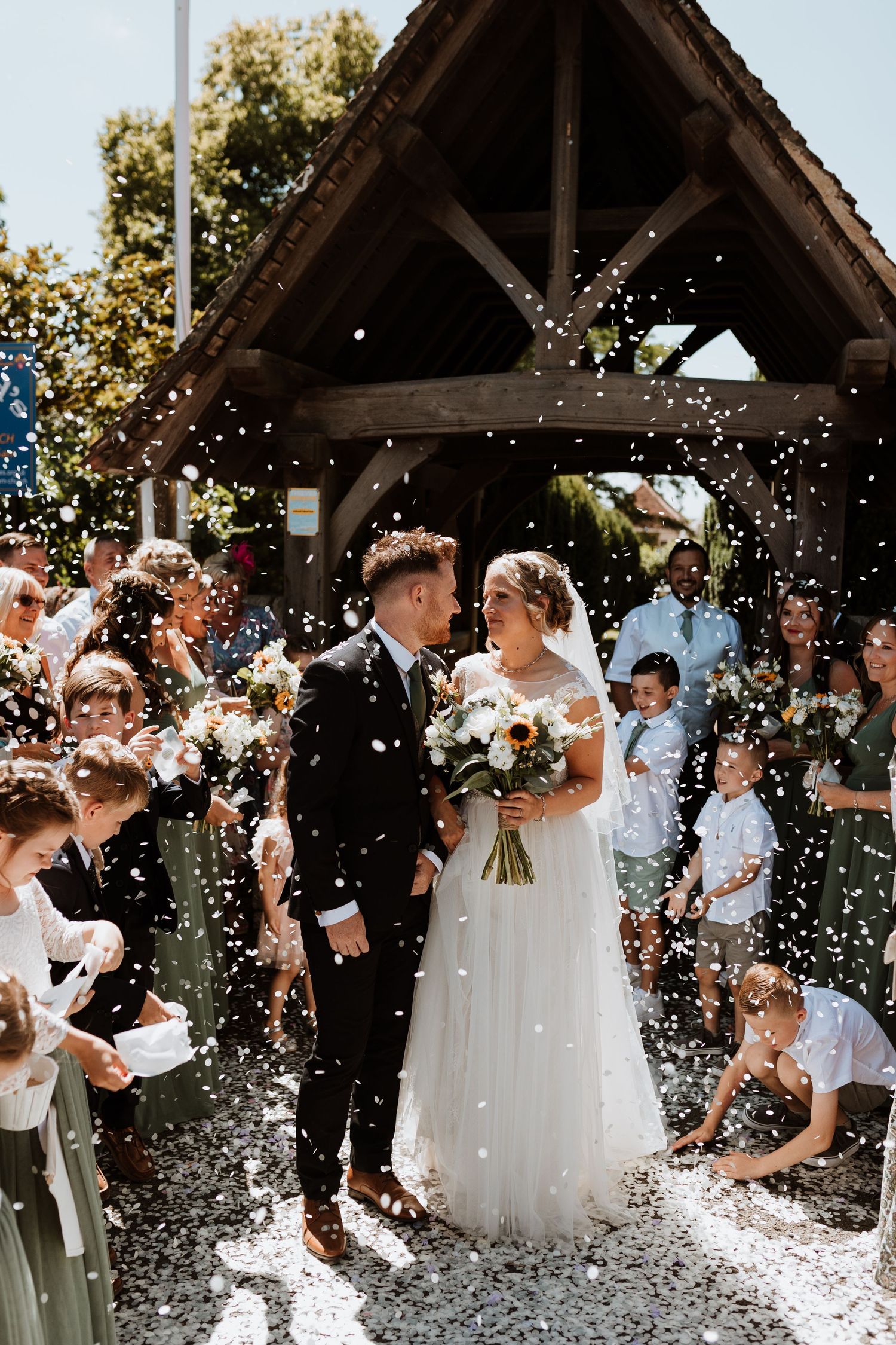 Wedding couple walks through a shower of confetti under a wooden gazebo while guests celebrate their special moment.
