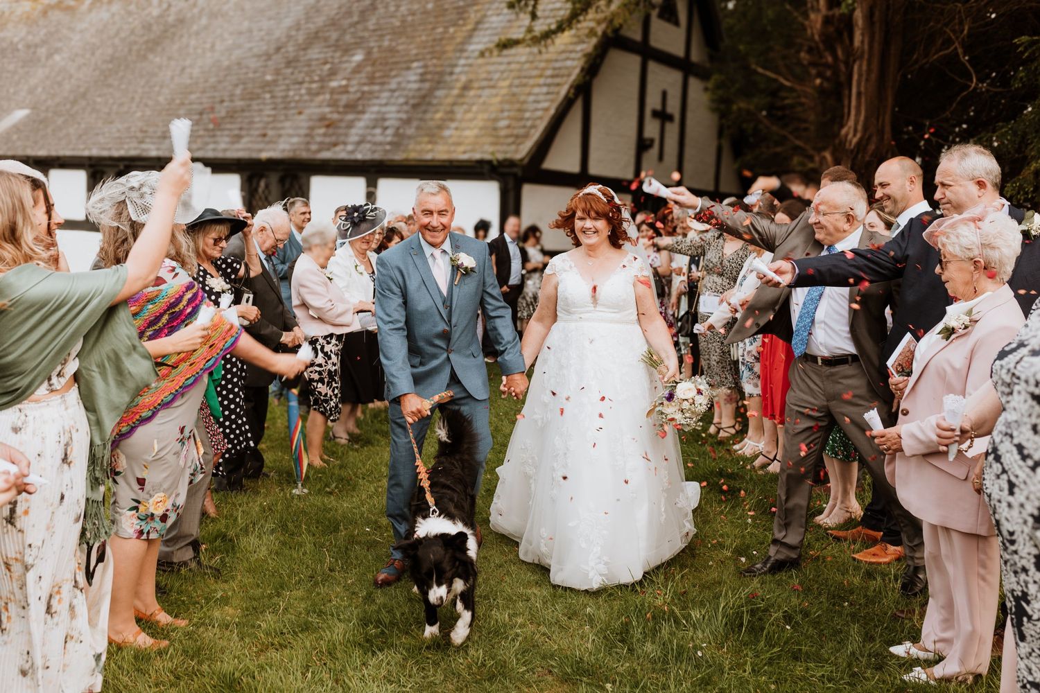 Wedding guests form a celebratory bubble-filled pathway on a lawn while joyful newlyweds walk under with their dog.