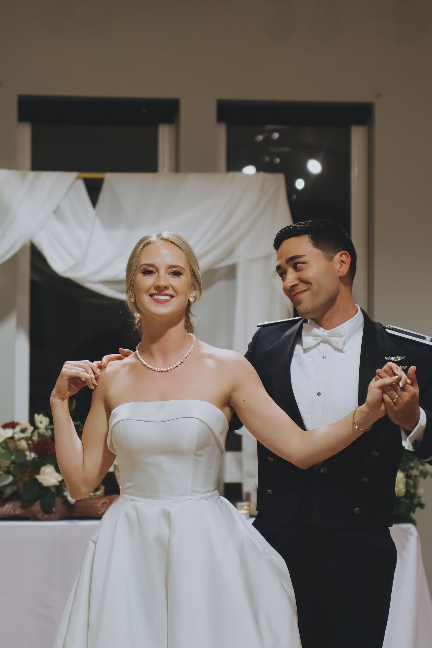 A couple shares their first dance at a wedding reception with white draping and subtle lighting in the background.