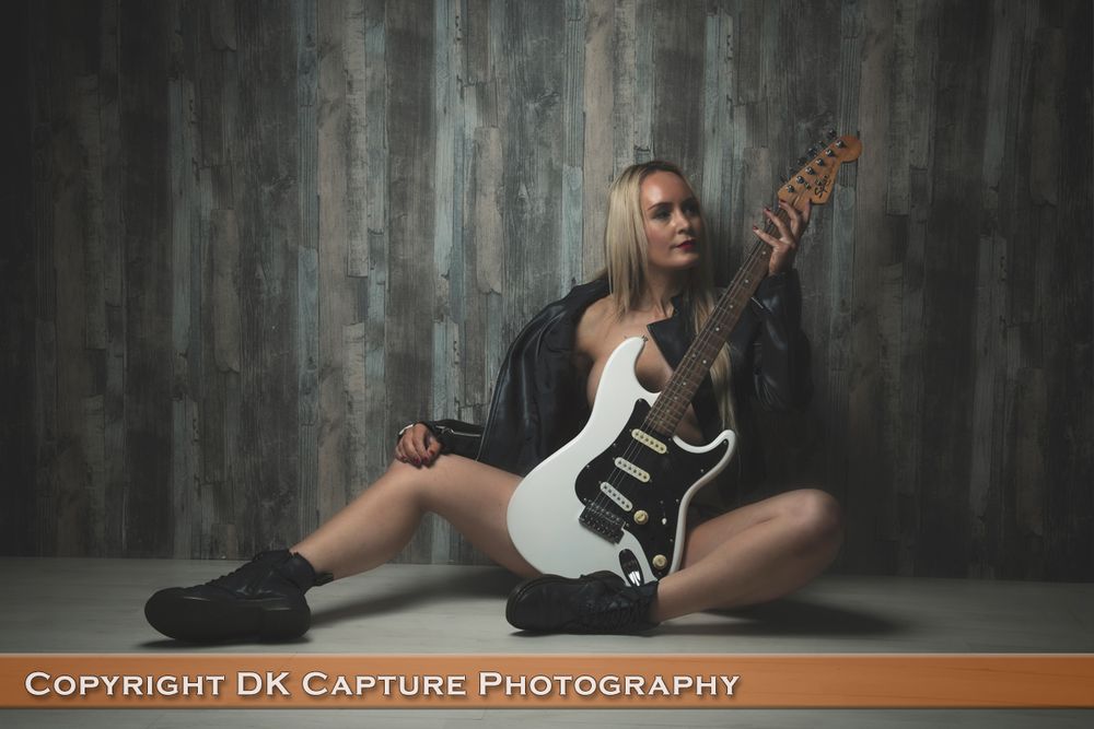 Musician poses with electric guitar while sitting on floor against textured gray wall in artistic portrait session.