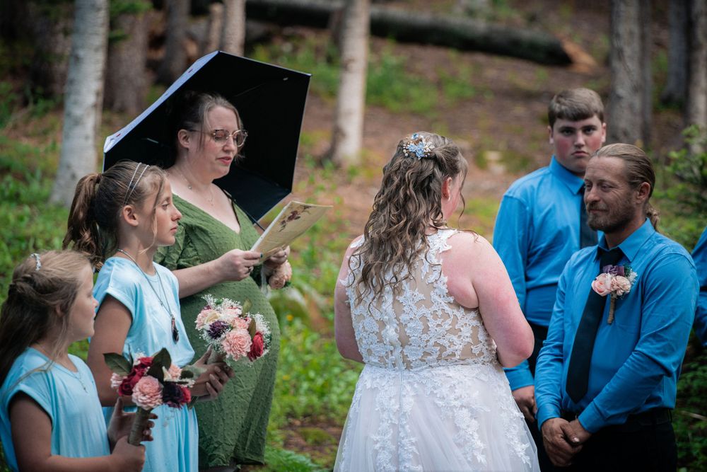 Forest wedding ceremony with attendants holding bouquets under a black umbrella.