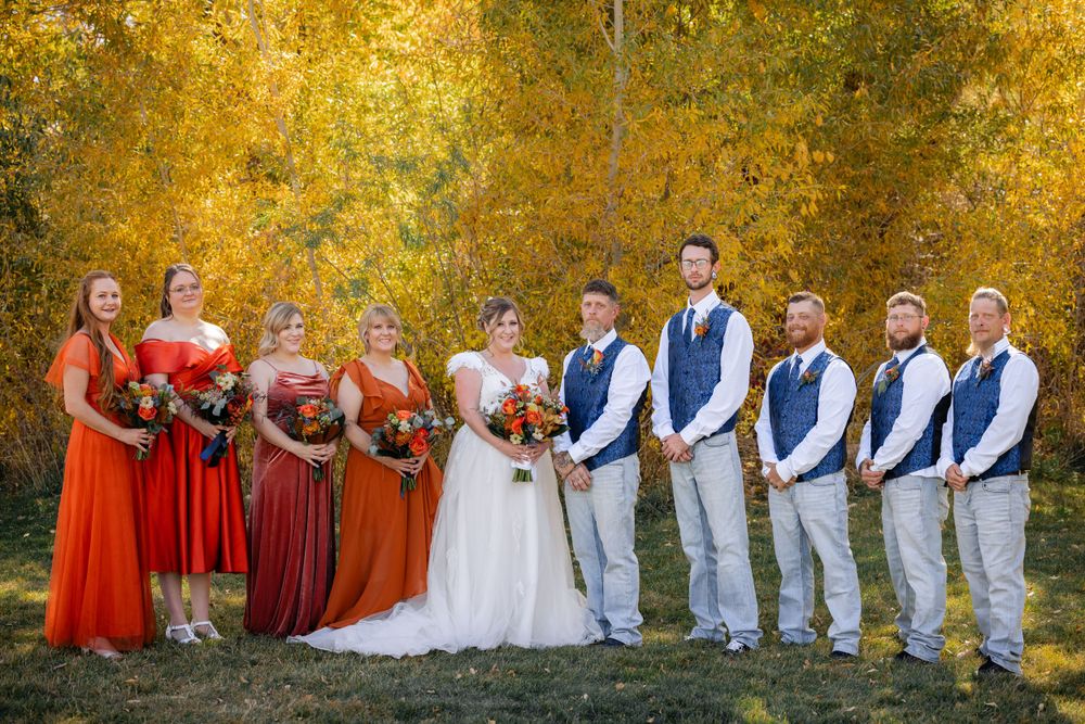 Wedding party in navy vests and orange dresses pose together against golden fall foliage.