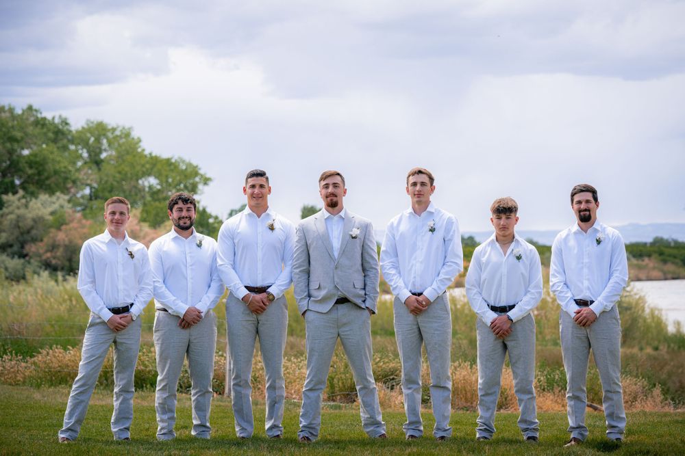 Wedding party in coordinated white shirts and gray pants poses on a grassy field.