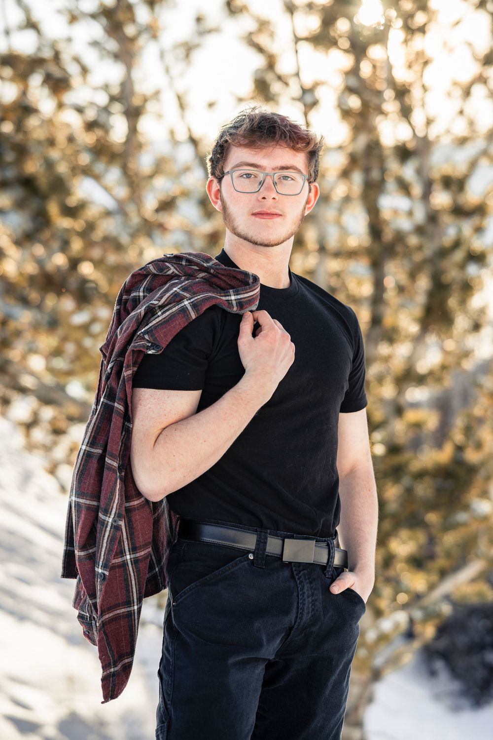 Young person in glasses wearing black t-shirt and holding plaid flannel shirt poses outdoors in snowy winter setting.