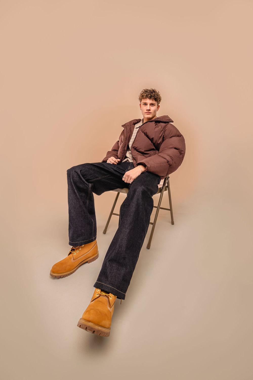 Person in brown puffer jacket and wide-leg jeans sits casually on folding chair wearing tan work boots against neutral backdrop.