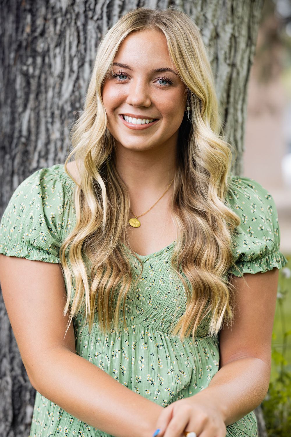 Portrait of someone in a mint green floral dress with long wavy blonde hair standing against a tree trunk.
