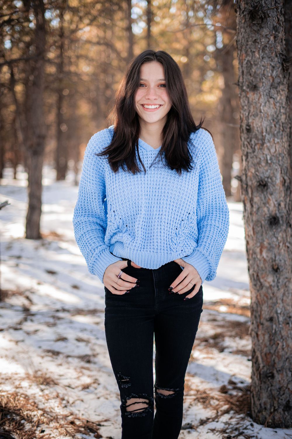 Winter portrait in blue sweater and ripped jeans against snowy forest backdrop.