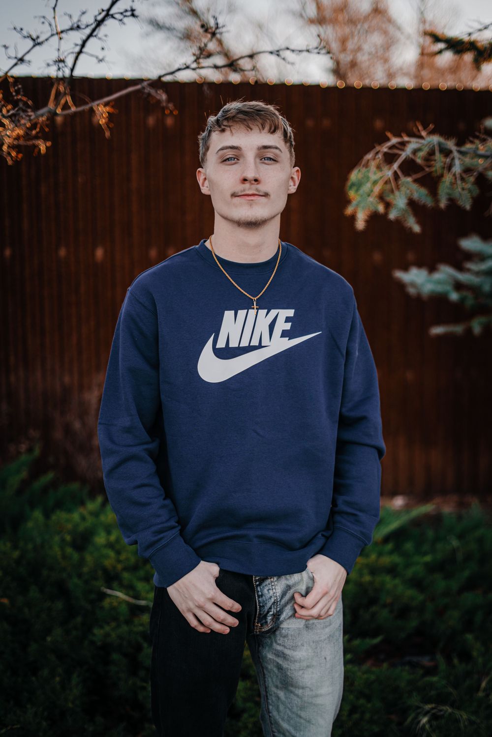 Person wearing navy blue Nike sweatshirt and jeans standing against wooden fence background.