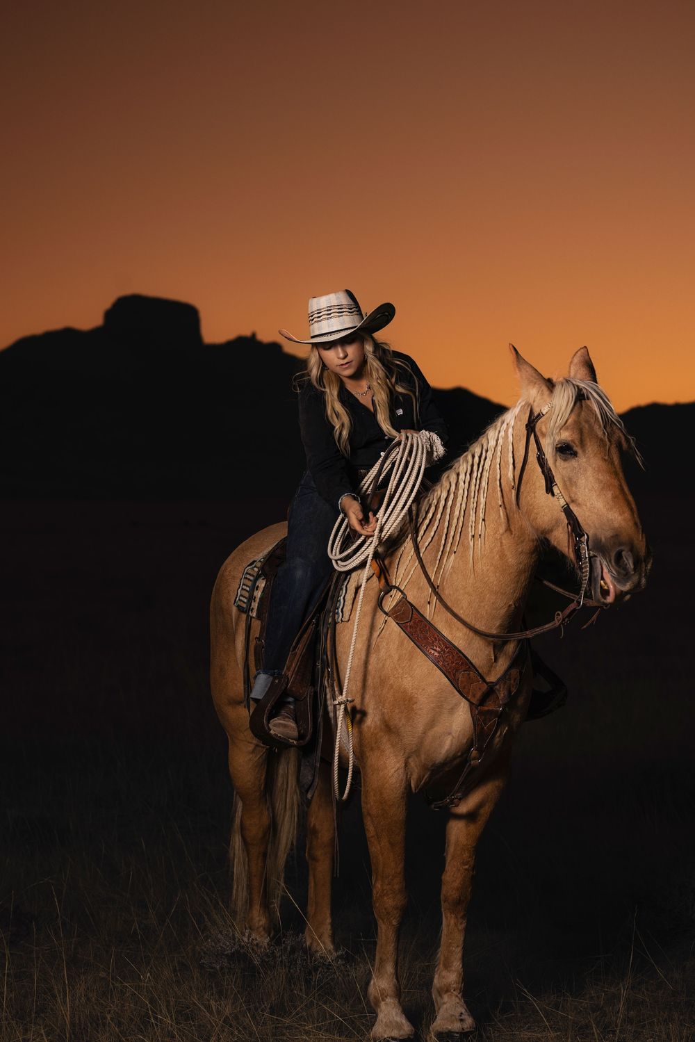 A person in cowboy hat rides a palomino horse against a dramatic orange sunset sky in western-style silhouette.