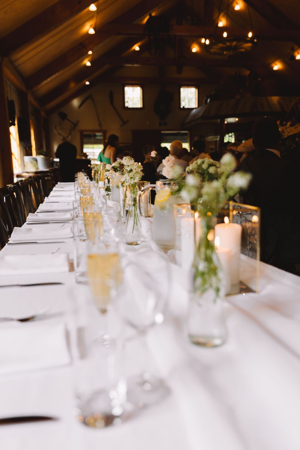 Elegant wedding reception table with glowing candles and white linens in rustic wooden venue.