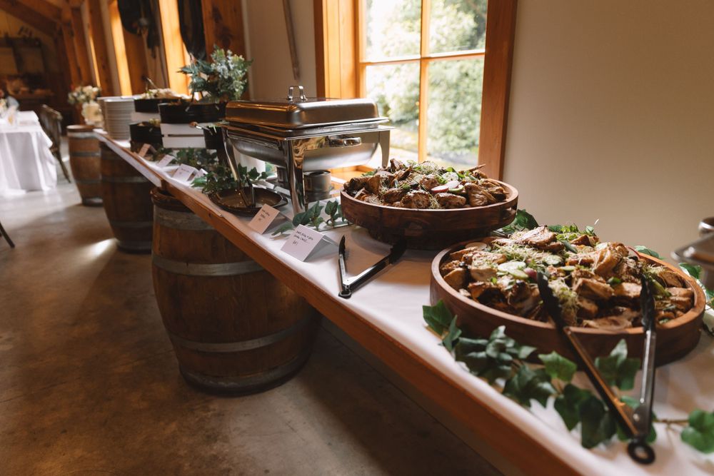 Rustic buffet setup with food warmers and decorative greenery on wooden barrels.