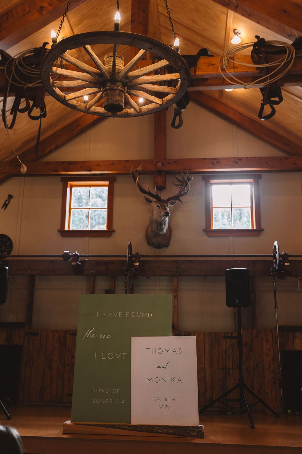 Rustic wedding venue interior with wagon wheel chandelier and mounted deer head between two windows.