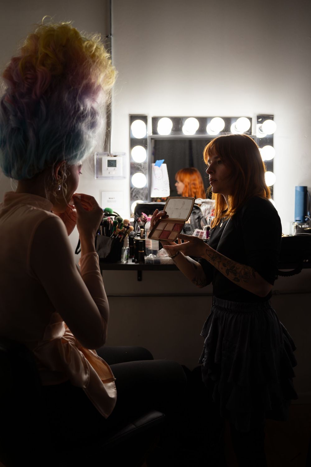 Dramatic backstage lighting captures makeup application in dressing room mirror.