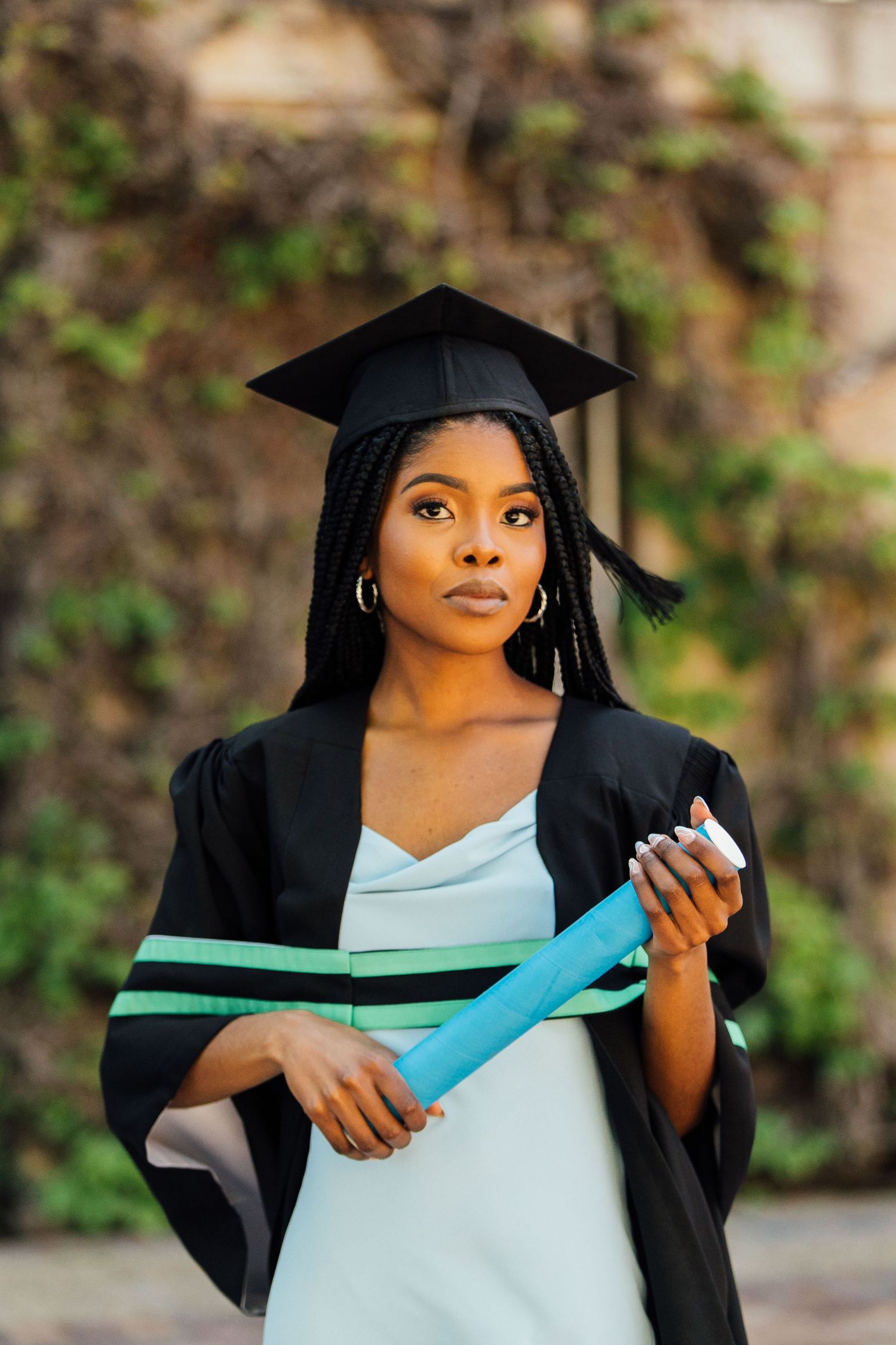 Graduate in black cap and gown holding turquoise diploma during outdoor graduation photo session.