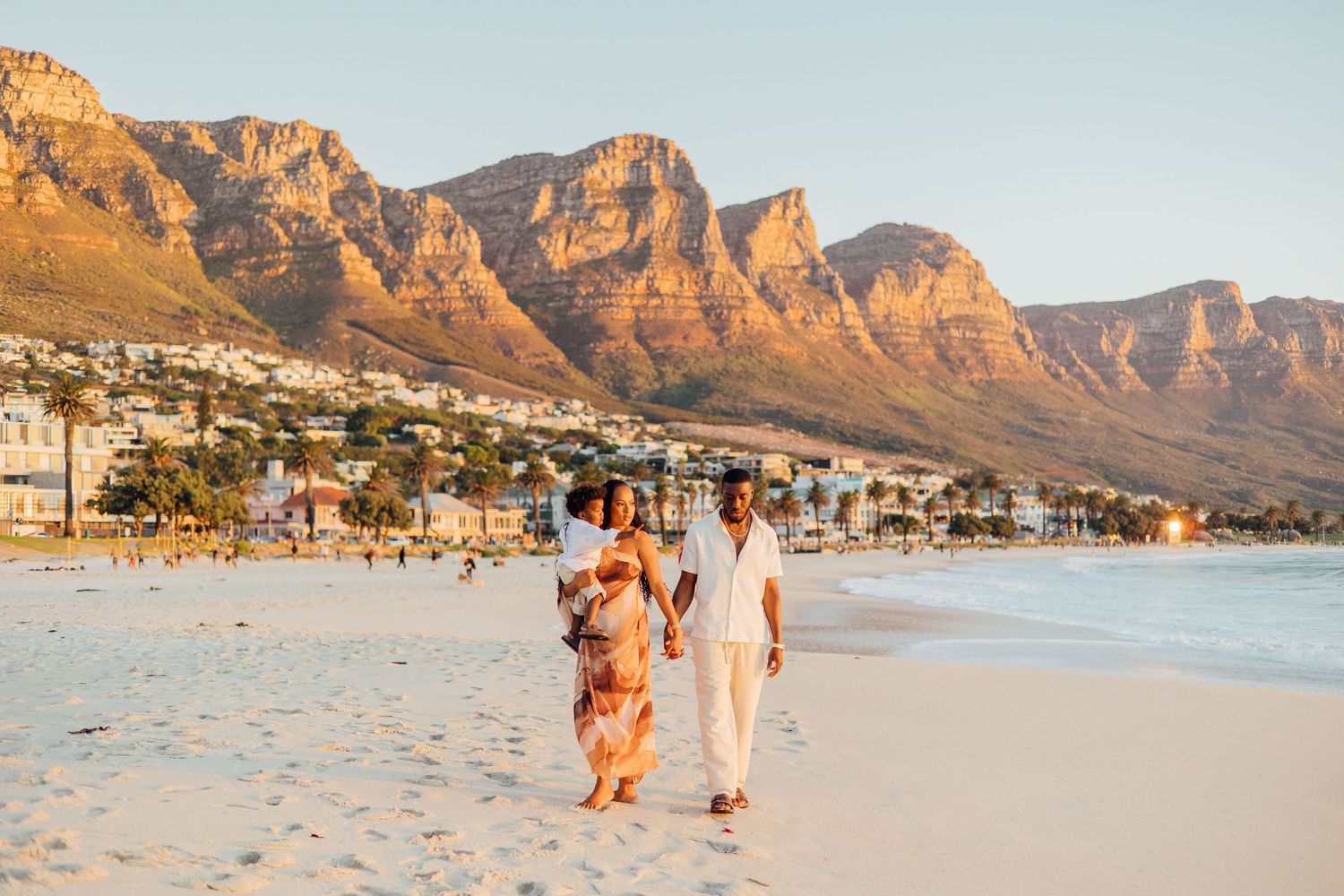 Two people stroll along Camps Bay Beach at sunset with the Twelve Apostles mountain range in Cape Town, South Africa.