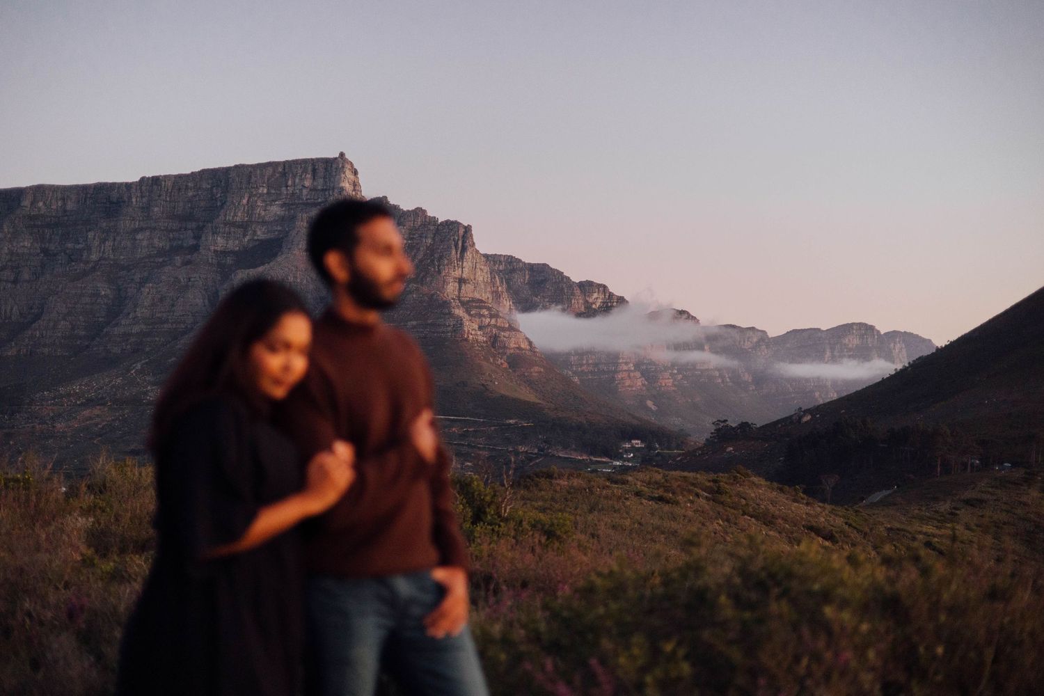 Two people embracing while hiking at sunset with Table Mountain's rugged cliffs and misty peaks in the background.