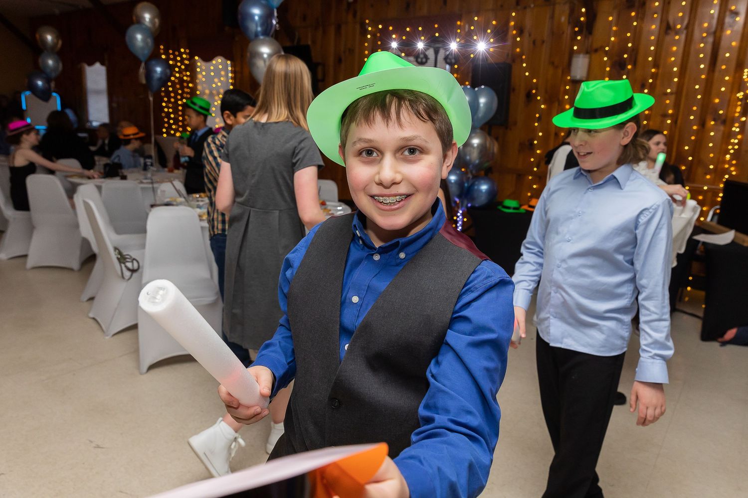 A young person in a vest and blue shirt celebrates at a party with festive lighting in the background.