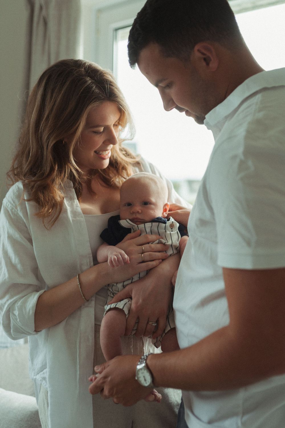 Parents in white shirts share a tender moment with their baby in a sun-lit room by a window.