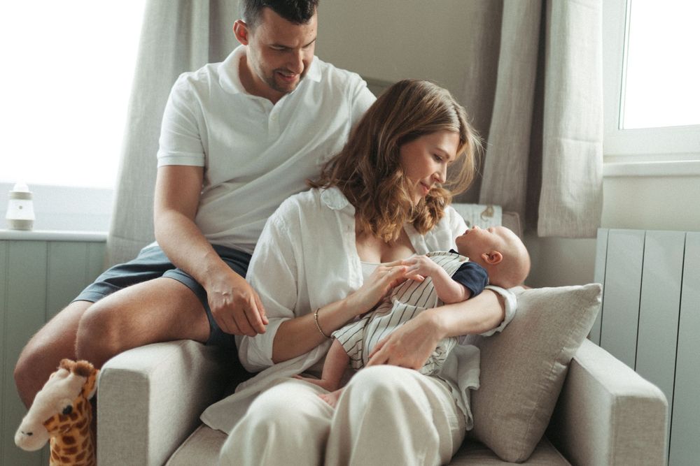 Family sitting together on a grey couch at home while bottle feeding their newborn baby.