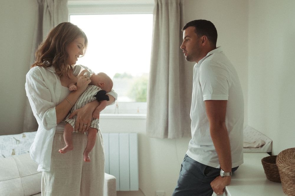 Two people in white clothing tenderly hold and look at a newborn baby near a bright window in a neutral-toned room.