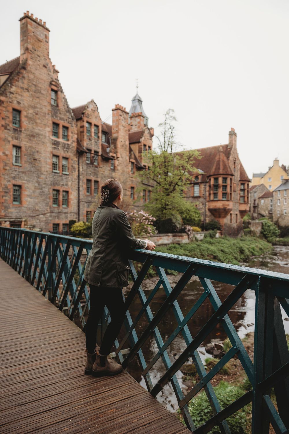 A figure stands on a wooden footbridge overlooking historic stone buildings in Dean Village, Edinburgh, Scotland.