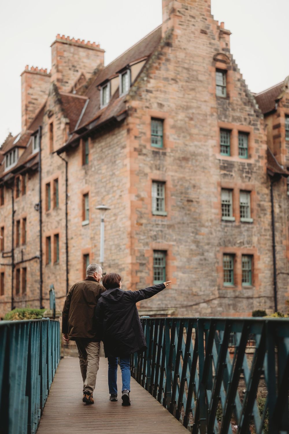 A couple walks together on a footbridge in front of historic stone buildings in Edinburgh, Scotland.