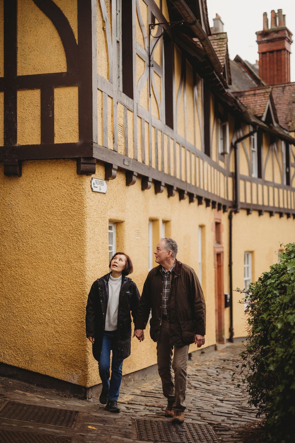 A couple walking hand in hand along a cobblestone path beside historic yellow Tudor-style buildings.